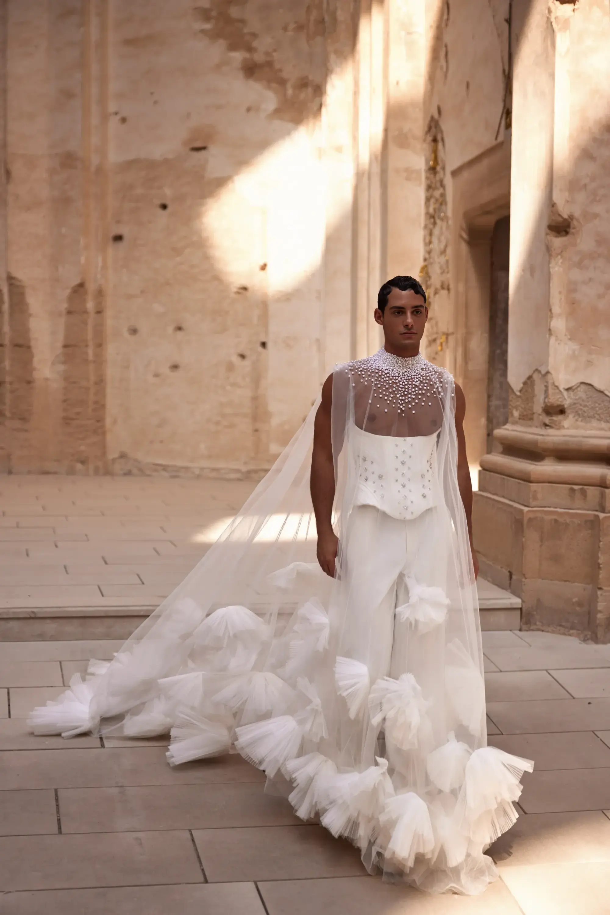 Tül detaylı uzun duvaklı ve işlemeli üst kısımlı avant-garde erkek gelinlik tasarımıyla moda podyumunda poz veren model; yüksek moda ve couture gelinlik trendleri.