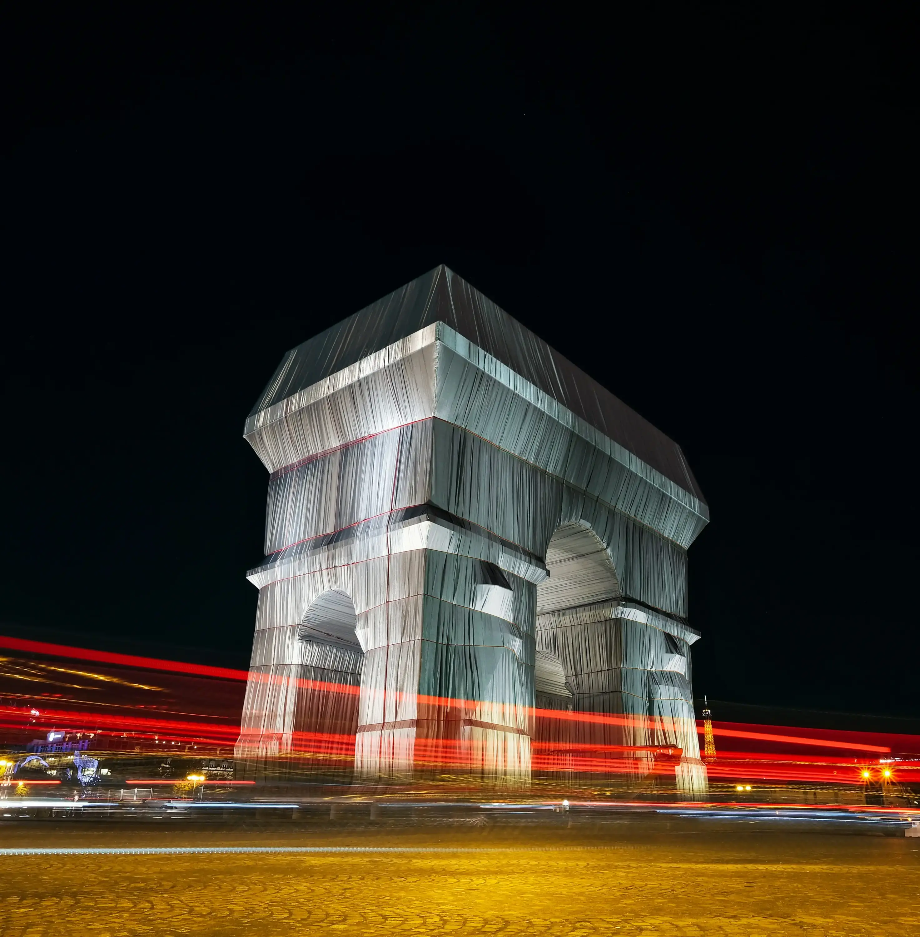 Christo ve Jeanne-Claude’un ‘Wrapped Arc de Triomphe’ enstalasyonunun gece çekimi, ışık izleriyle Paris’te çağdaş sanat görünümü.