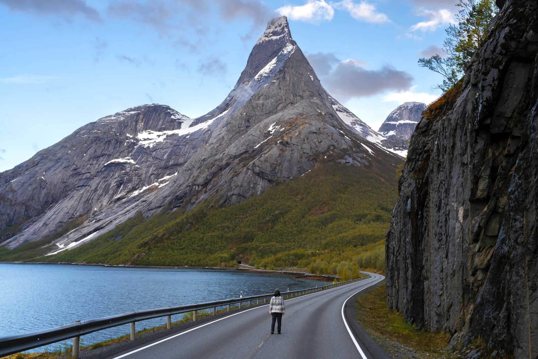 Fiyord kıyısında kıvrılan yolda yürüyen kişi ve arka planda yükselen dramatik, karlı Norveç dağlarıyla manzara fotoğrafı.