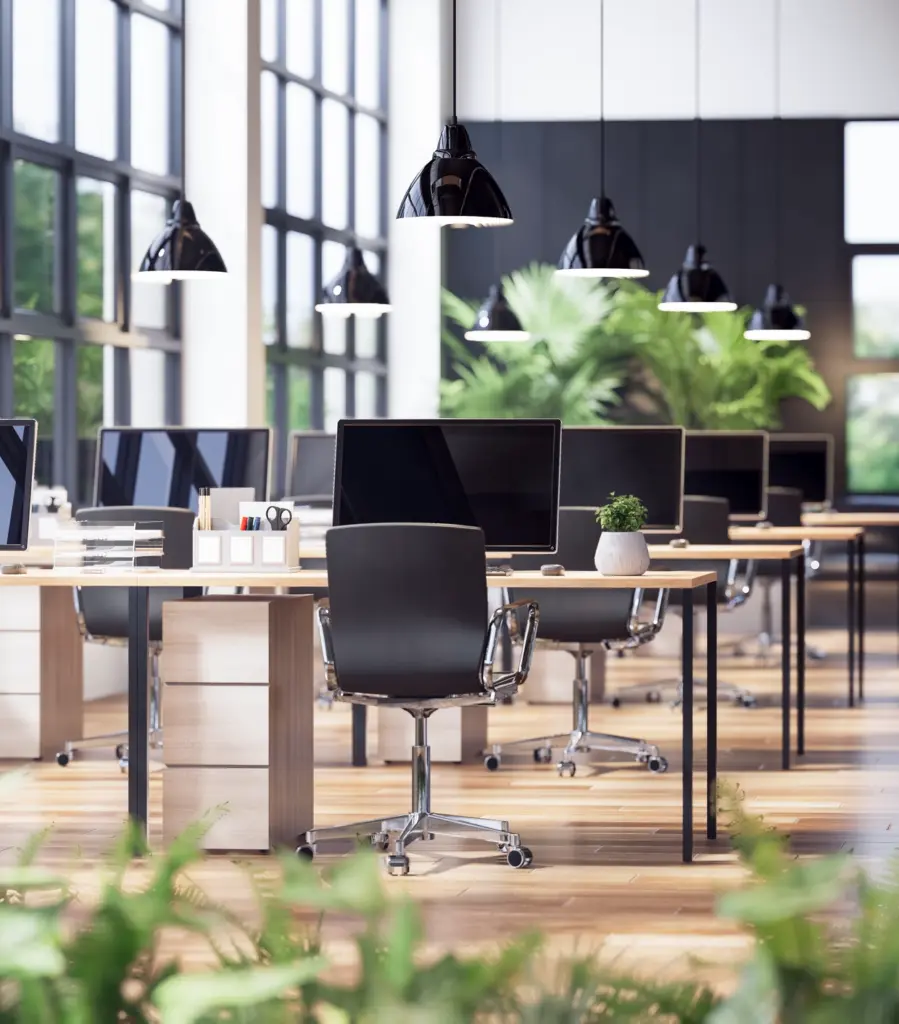 Geniş pencereli, doğal ışık alan modern ofis alanında sıralanmış bilgisayarlar, ergonomik çalışma koltukları ve yeşil bitkilerle tasarlanmış sürdürülebilir açık ofis ortamı.