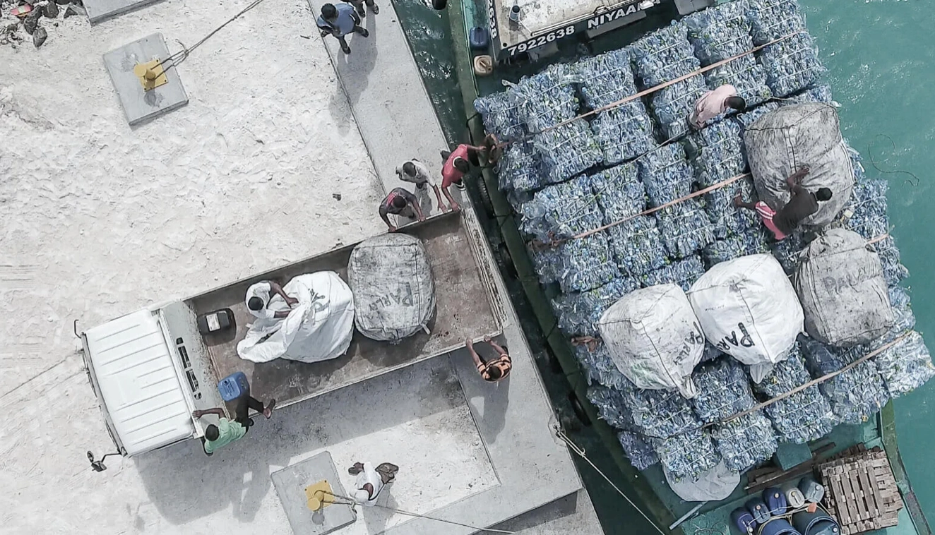 Okyanus plastiklerinin toplanıp geri dönüşüm merkezine taşındığı süreçten bir hava çekimi; tekneden kamyona aktarılan sıkıştırılmış plastik şişeler ve çalışan ekip.