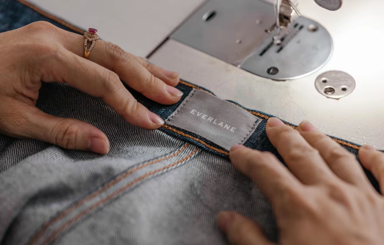Everlane denim ürününün etiketi dikilirken ellerin yakın plan görüntüsü, etik üretim ve sürdürülebilir moda vurgusu.