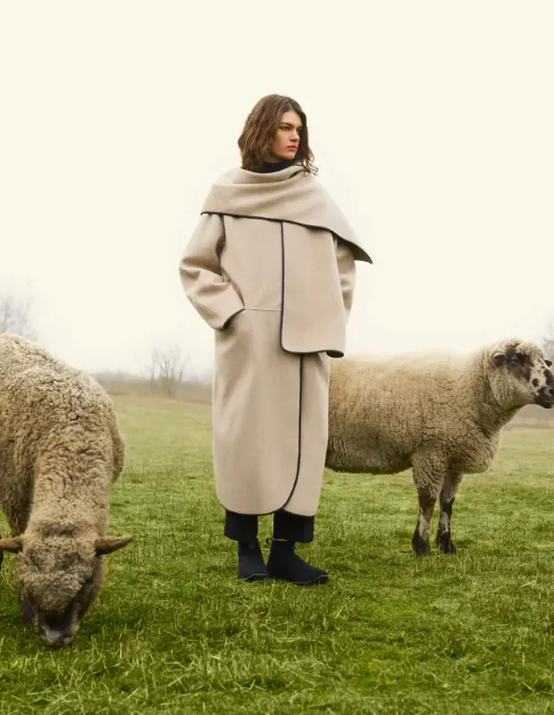 Doğal yün üretimi ve sürdürülebilir moda temasını yansıtan fotoğraf: Bej tonlarında oversized bir kaban giyen model, sisli bir kır manzarasında otlayan iki koyunun arasında duruyor; doğal malzeme, etik üretim ve çevre dostu tasarım vurgusu taşıyan editorial çekim.