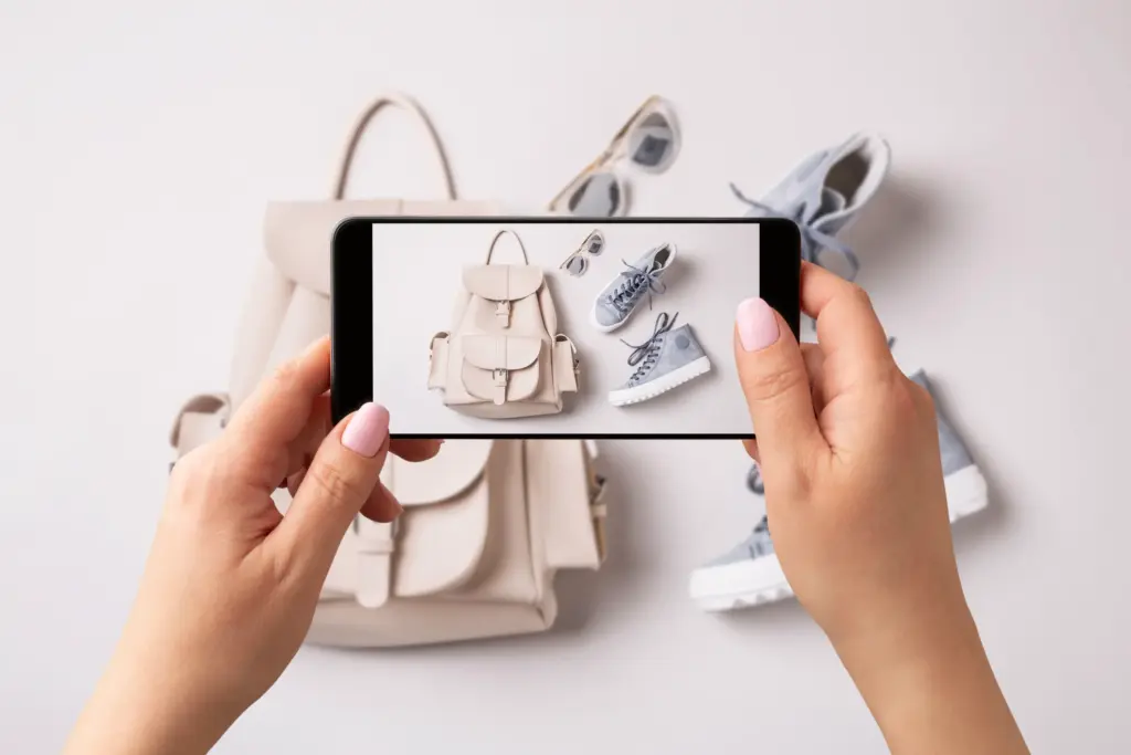 Bir kişinin akıllı telefonla krem rengi sırt çantası ve mavi spor ayakkabı kombinasyonunun fotoğrafını çektiği ikinci el moda ve dijital alışveriş temalı görsel.
