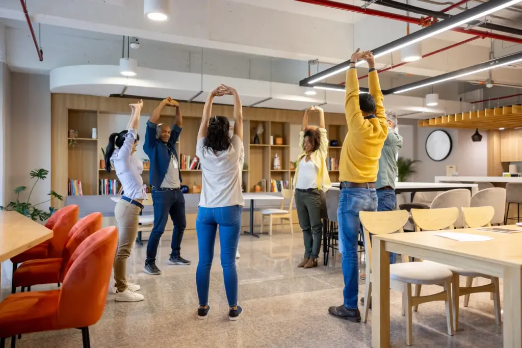 Ofis ortamında çalışanların toplu esneme egzersizi yaptığı, kurumsal wellbeing uygulamalarını temsil eden modern işyeri sahnesi.