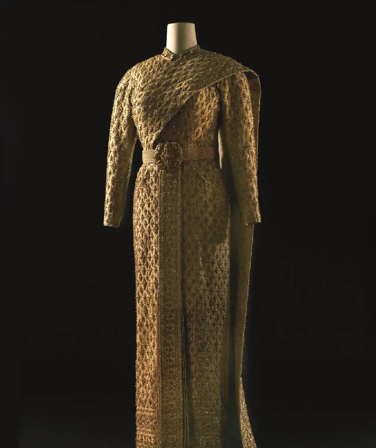 Altın tonlarında, yoğun işlemeli ve kemerli uzun couture elbise; müze sergilemesinde dramatik ışık altında sunulan zamansız haute couture tasarımı.