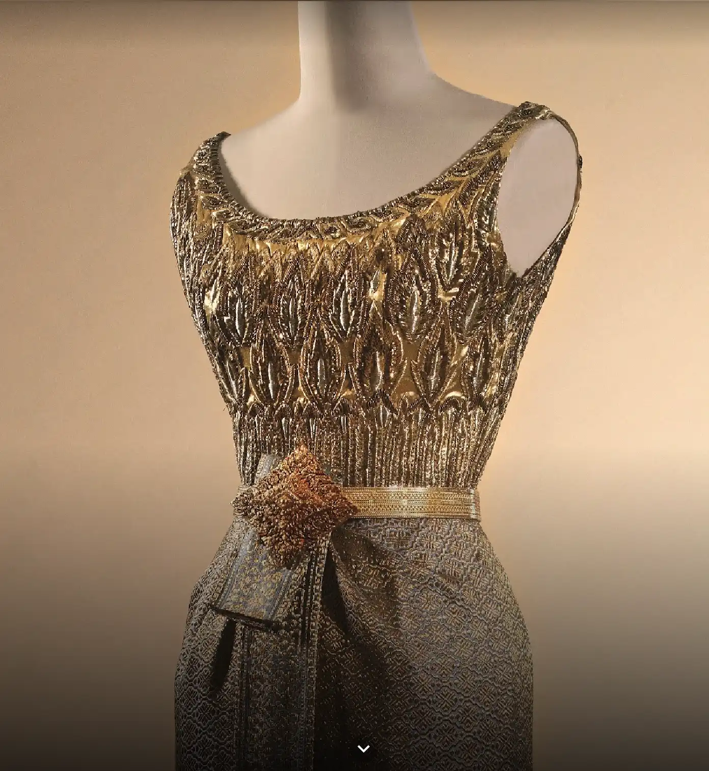 Altın iplik ve boncuklarla işlenmiş, bel kısmında heykelsi detay bulunan couture elbise; haute couture işçiliğini öne çıkaran yakın plan sergi görünümü.