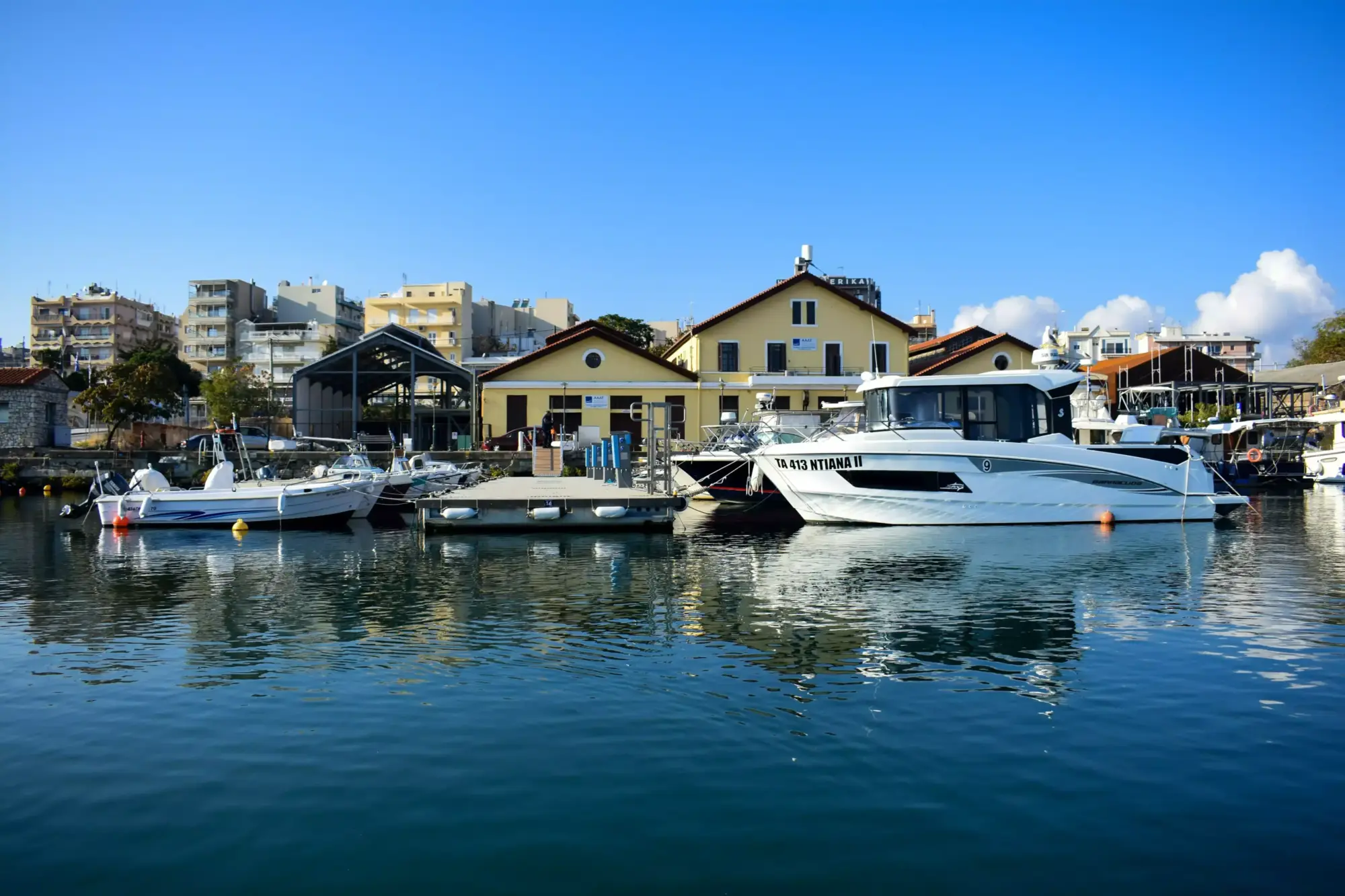 Alexandroupoli limanında, masmavi berrak sular üzerinde demirlemiş, farklı boyutlarda modern beyaz yatlar ve teknelerin sıralandığı, arkasında ise sarı duvarlı ve kiremit çatılı geleneksel görünümlü binalar ile çok katlı apartmanların göründüğü güneşli ve sakin bir gün manzarası.