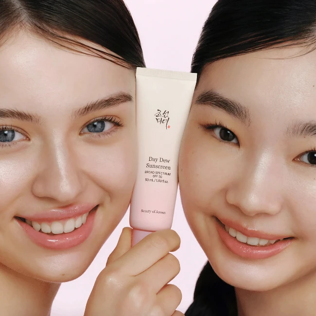 Farklı cilt tonlarına sahip, parlak gülümsemeleri ve sağlıklı görünen ciltleriyle iki kadın, ortalarında Beauty of Joseon Day Dew Sunscreen SPF 50 güneş kremi tüpünü tutarak, cildin güneşten korunmasının ve nemlenmesinin önemini doğal bir yaşam tarzı sunumuyla vurguluyor.