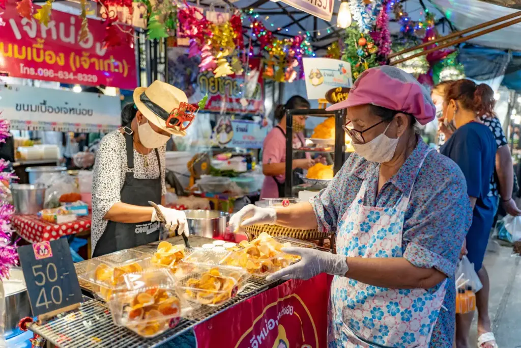 Phuket'in bohem atmosferli Chillva Pazarı'nda maske takan iki satıcı, renkli süslemelerle donatılmış tezgahlarında Tayland'ın yerel sokak lezzetlerini özenle hazırlayıp plastik kaplarda müşterilere sunuyor, fiyat tabelası da ürün çeşitliliğini gösteriyor.