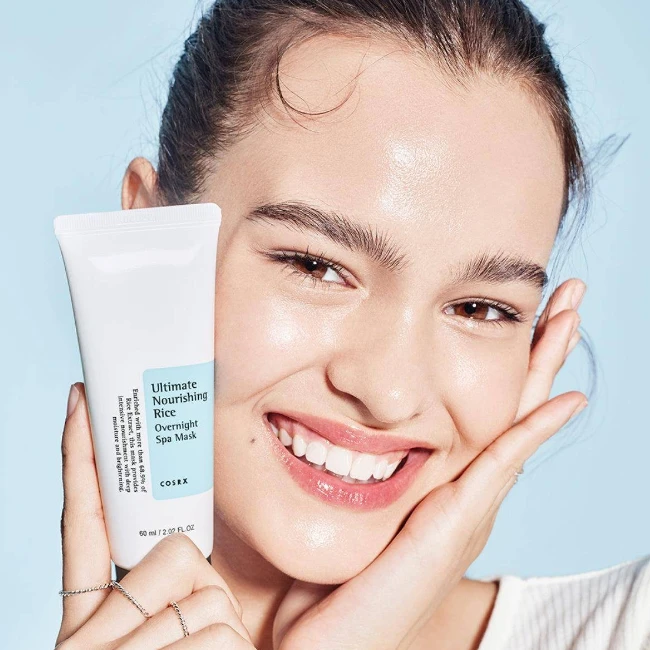 Pürüzsüz ve sağlıklı görünen cildiyle samimi bir gülümseme sergileyen genç bir kadın, elinde cilt bariyerini güçlendiren ve yoğun nem sağlayan Cosrx Ultimate Nourishing Rice Overnight Spa Mask ürününü tutuyor.
