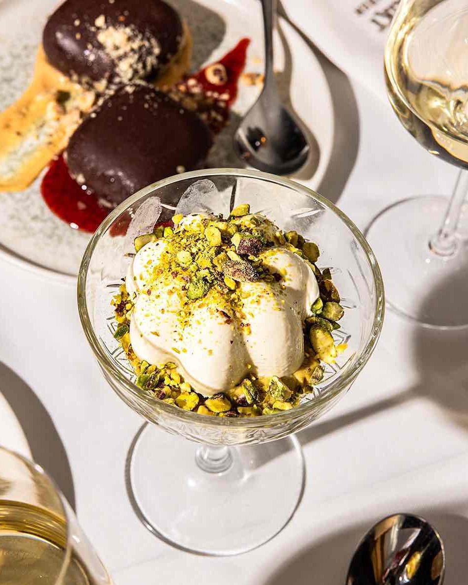 Üsküp seyahat rehberindeki gastronomi önerilerinden biri olan Distrikt Bar & Kitchen'da sunulan, ince çekilmiş Antep fıstıklarıyla süslenmiş kremalı bir tatlı ile arka planda görünen çikolatalı hamur işleri ve kadeh şarap içeren şık bir masa detayı.