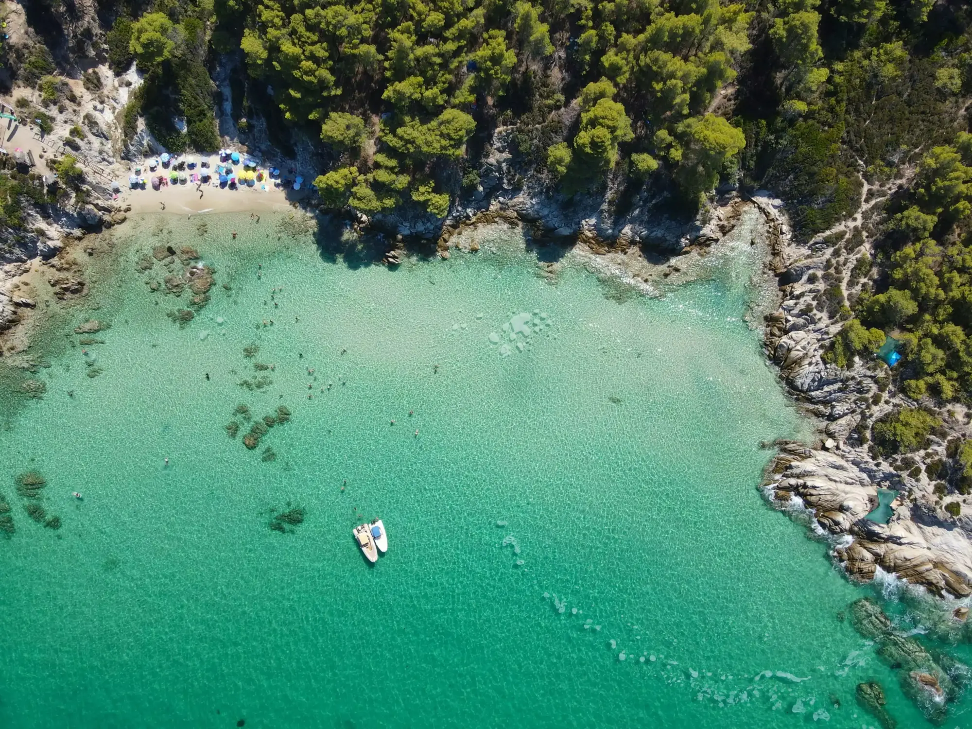 Halkidiki'deki çam ağaçlarıyla kaplı tepelerin çevrelediği, turkuaz renkli berrak denizi ve renkli şemsiyelerle dolu kumsalıyla dikkat çeken güzel bir koyun havadan görünümü, iki küçük teknenin ve yüzen insanların olduğu huzurlu bir yaz manzarasını sergiliyor.