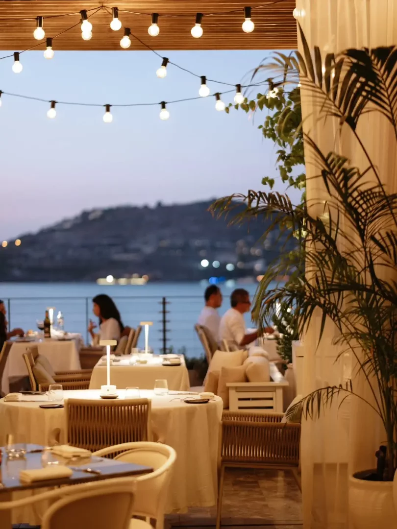 Akşamüstü, Bodrum'da deniz kenarında, sarkaç lambaların ışıl ışıl aydınlattığı şık bir açık hava restoranında, beyaz örtülü masalarda yemek yiyen misafirler huzurlu bir ambiyansın tadını çıkarıyor.