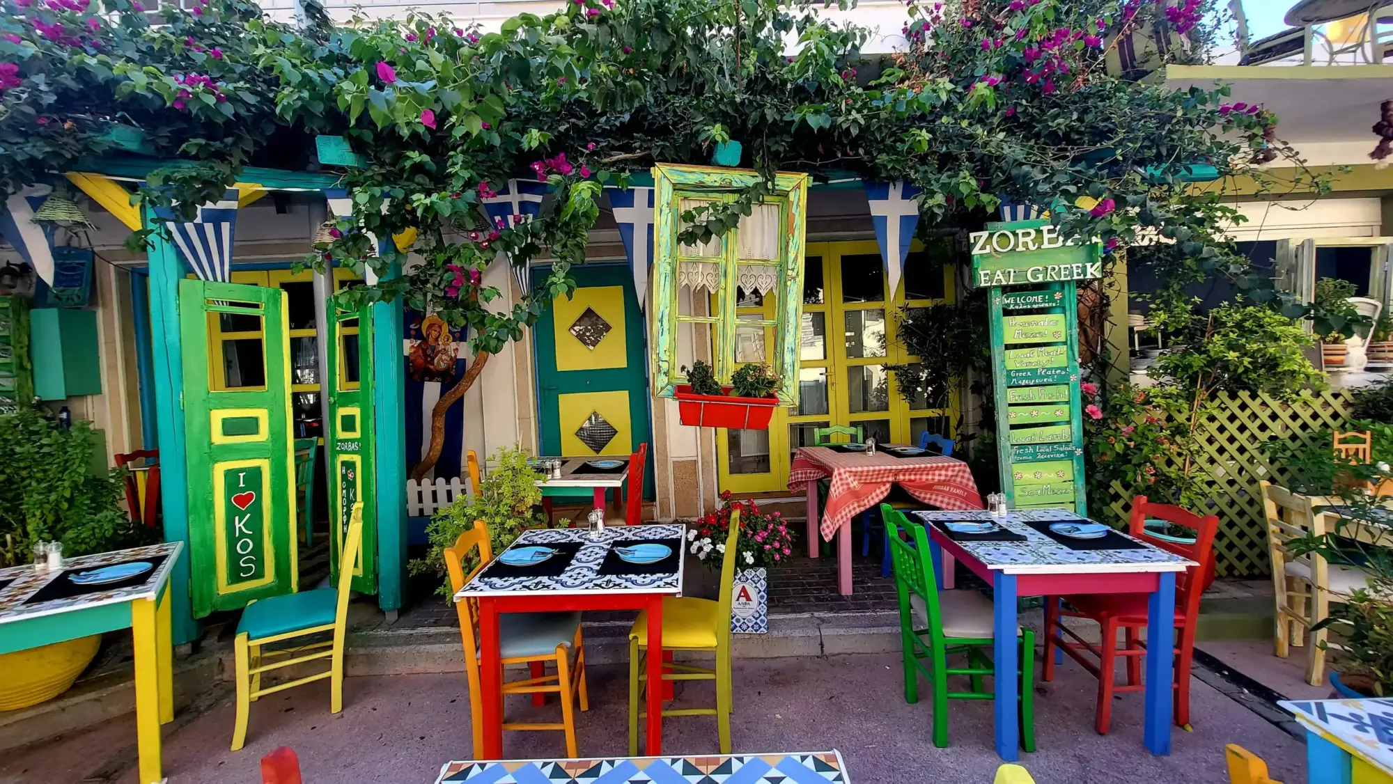 Kos adasındaki canlı ve rengarenk bir Yunan restoranının dış mekanı, pembe begonvillerle süslü yeşil bitkilerin çevrelediği parlak sarı ve yeşil kapıları, sallanan mavi-beyaz Yunan bayrakları ve çeşitli renklerdeki sandalyelerle dolu açık hava masalarını gösteriyor, neşeli bir yemek deneyimi vaat eden davetkar bir atmosfer sunuyor.