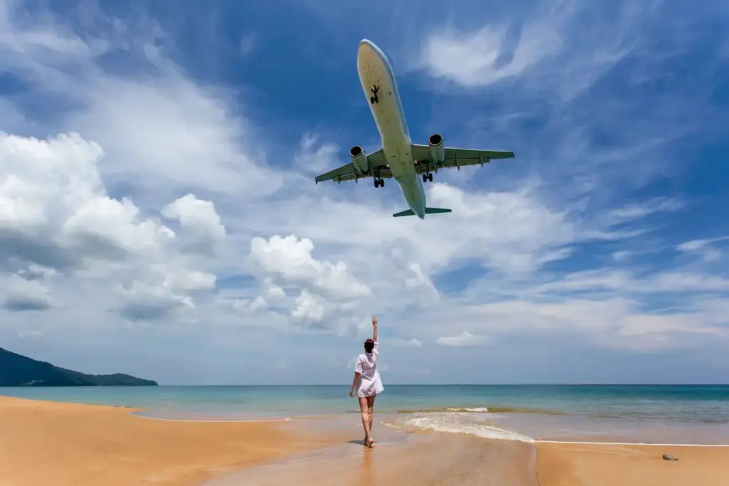 Phuket'in kuzeyindeki ikonik Mai Khao Plajı'nda, bir kişinin turkuaz kıyılara doğru dönerek başının sadece birkaç metre üzerinden alçalmakta olan devasa bir yolcu uçağına el salladığı, mavi gökyüzünde beyaz bulutların eşlik ettiği, adanın en dikkat çekici gezilecek yerlerinden birini yansıtan canlı bir an.