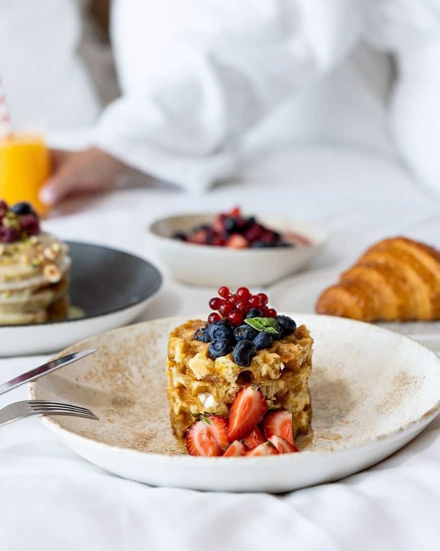 Üzeri çilek, yaban mersini ve frenk üzümü gibi taze orman meyveleriyle özenle süslenmiş, parlak şurupla gezdirilmiş waffle veya pancake kulesinin ön planda olduğu bu görüntü, arka planda bulanık görünen kruvasan, meyve kasesi ve portakal suyuyla birlikte, Üsküp seyahat rehberinde bahsedilen lüks bir şehir otelindeki konforlu kahvaltı deneyimini gözler önüne seriyor.