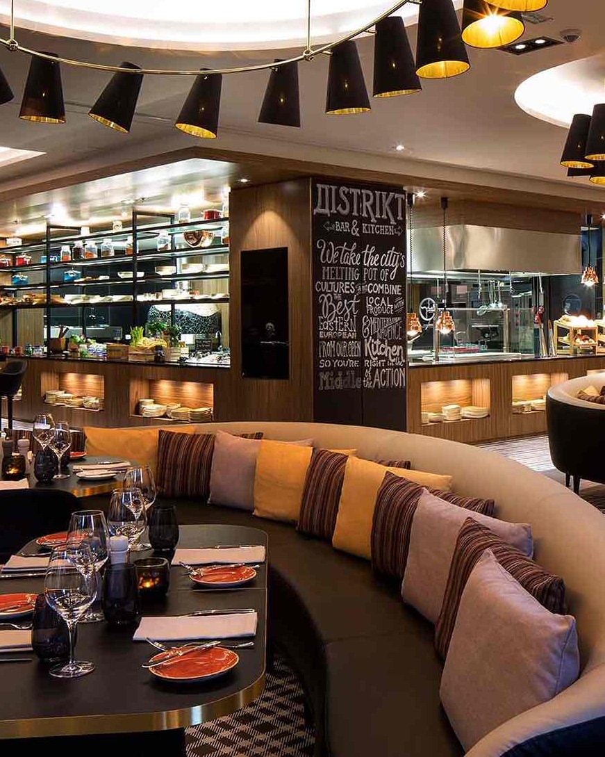 Modern konik siyah lambaların aydınlattığı, şık kavisli banklara dizilmiş renkli yastıkların bulunduğu ve masaların yemek servisine hazırlandığı Üsküp'teki Distrikt Bar & Kitchen'ın açık mutfaklı zarif iç mekanı.