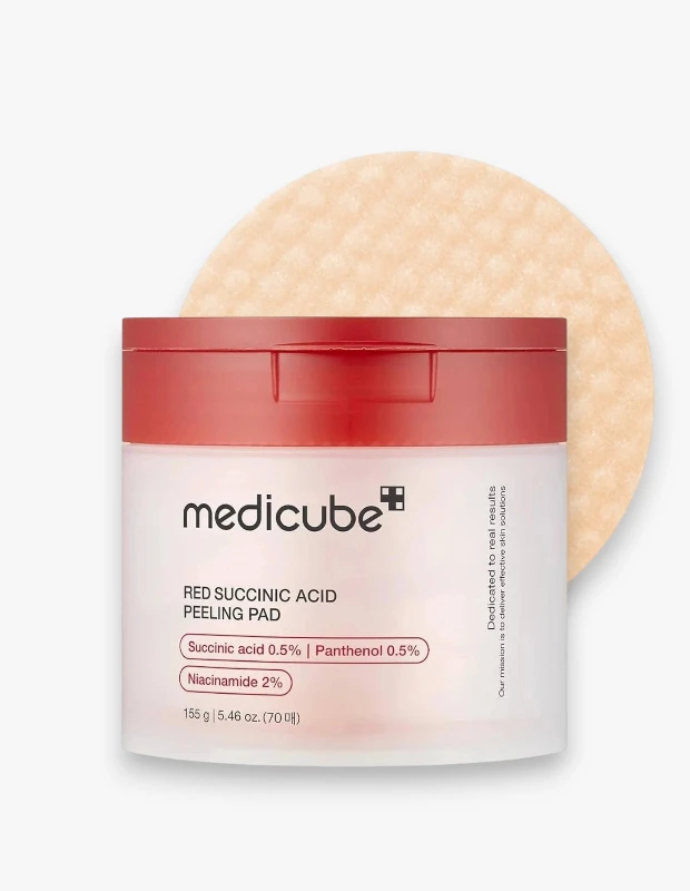Beyaz zemin üzerinde, kırmızı kapaklı mat beyaz bir Medicube RED SUCCINIC ACID PEELING PAD kavanozu ile arkasından görünen, hafif bej renkli ve dokulu bir peeling pedi sergileniyor; ürün üzerinde %0.5 Süksinik Asit, %0.5 Panthenol ve %2 Niasinamid içerdiği yazılı.