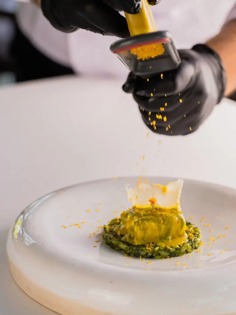 Mezra Yalıkavak restoranında bir şefin siyah eldivenli elleri, parlak sarı soslu ve çıtır garnitürlü yeşil tabanlı ana yemeğin üzerine sarı pulcuklar serpiştirerek yemeğin sunumuna son dokunuşları yapıyor.
