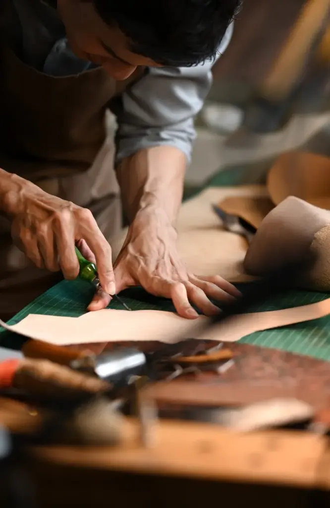 El işçiliğiyle deri kesen zanaatkârın atölye ortamında üretim sürecini gösteren detaylı craft moda görseli.