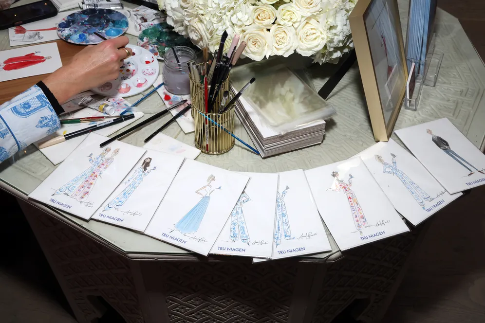 Moda tasarım sürecini gösteren bir çalışma masasında yayılan el çizimi moda eskizleri, boya paleti, fırçalar ve kalemlerle birlikte el işçiliği ve tasarım hikâyesinin yaratım sürecini yansıtan detaylı atölye sahnesi.