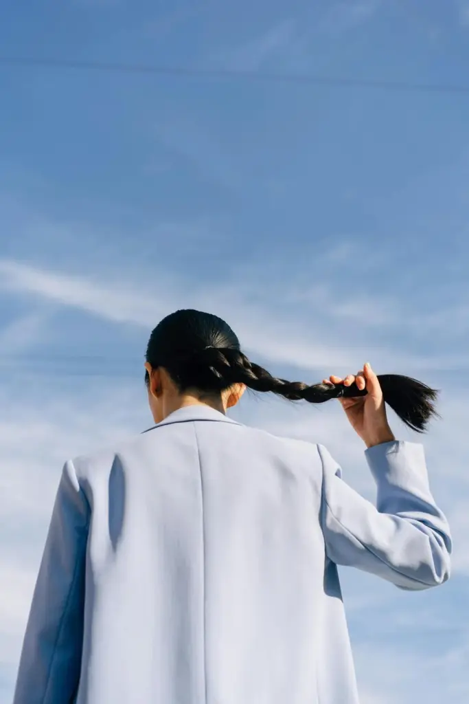 Açık mavi blazer ceket giyen bir kadının saçını örerken gökyüzü fonunda görüntülendiği modern moda karesi; sade silüetler ve doğal ışıkla öne çıkan çağdaş stil vurgusu.