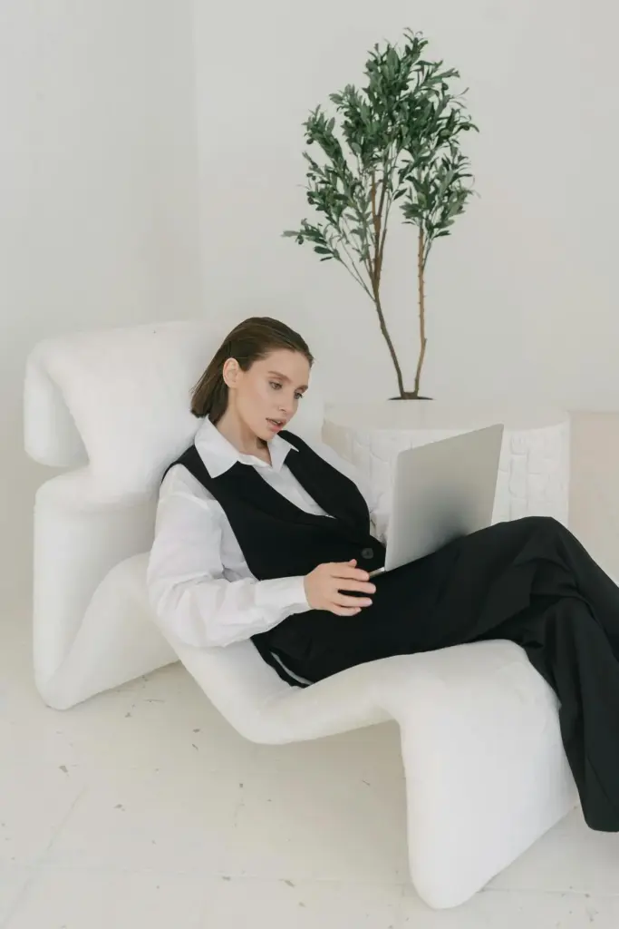 Beyaz ve siyah parçalarla rahat ama şık bir kombin giyen genç kadının laptopla çalıştığı modern iç mekân sahnesi; çağdaş ofis stili ve konfor odaklı moda yaklaşımı.
