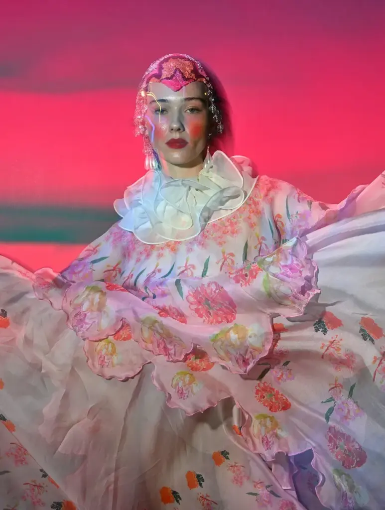 Pembe ve çiçek desenli dramatik elbise giyen model; sahne ışıkları altında avangart, teatral moda görünümü.