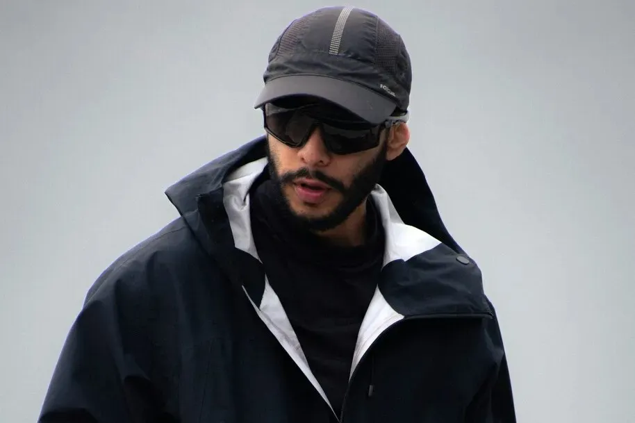 Outdoor şehir stilinde siyah mont, şapka ve güneş gözlüğü takan erkek; modern fonksiyonel moda trendlerini yansıtan sokak stili portresi.