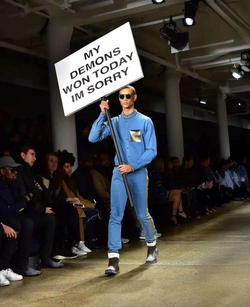 Bir moda defilesinde “My Demons Won Today I'm Sorry” yazılı pankart taşıyan modelin podyumda yürüdüğü provokatif moda sahnesi.