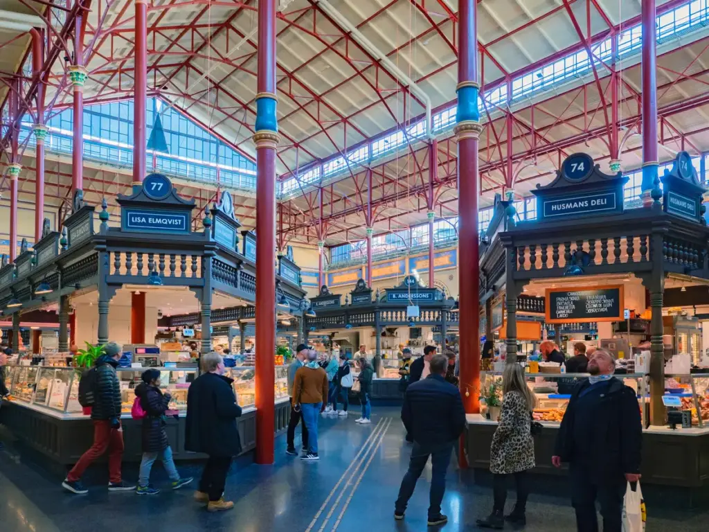 Östermalms Saluhall içinde, Stockholm'ün gezilecek yerlerinden biri olan tarihi gurme pazarında, yüksek tavanı destekleyen kırmızı ferforje sütunların ve cam panellerin aydınlattığı geniş salonda, müşteriler geleneksel ahşap tezgahların önünde şarküteri ve çeşitli lezzetleri inceliyor.