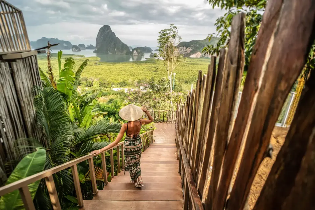 Konik şapkalı ve desenli elbiseli bir kadın, Samet Nangshe Viewpoint'ten ahşap bir merdivenden inerek, Phang Nga Körfezi'nin kireçtaşı adaları ve yemyeşil mangrov ormanlarıyla bezenmiş nefes kesici panoramik manzarasının tadını çıkarıyor, bu eşsiz görüntü Phuket'in yakınındaki turkuaz kıyılar rotalarında unutulmaz bir durak.