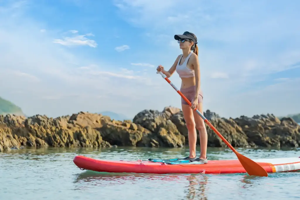 Phuket'in berrak turkuaz kıyılarında, güneşli ve açık bir havada, şapkalı ve güneş gözlüklü bir kadın, kırmızı stand-up paddleboard üzerinde denizin tadını çıkararak kürek çekiyor; arka planda adanın karakteristik kayalık oluşumları ve masmavi gökyüzü, su sporları ve huzurlu bir keşif için ideal ortamı gözler önüne seriyor.