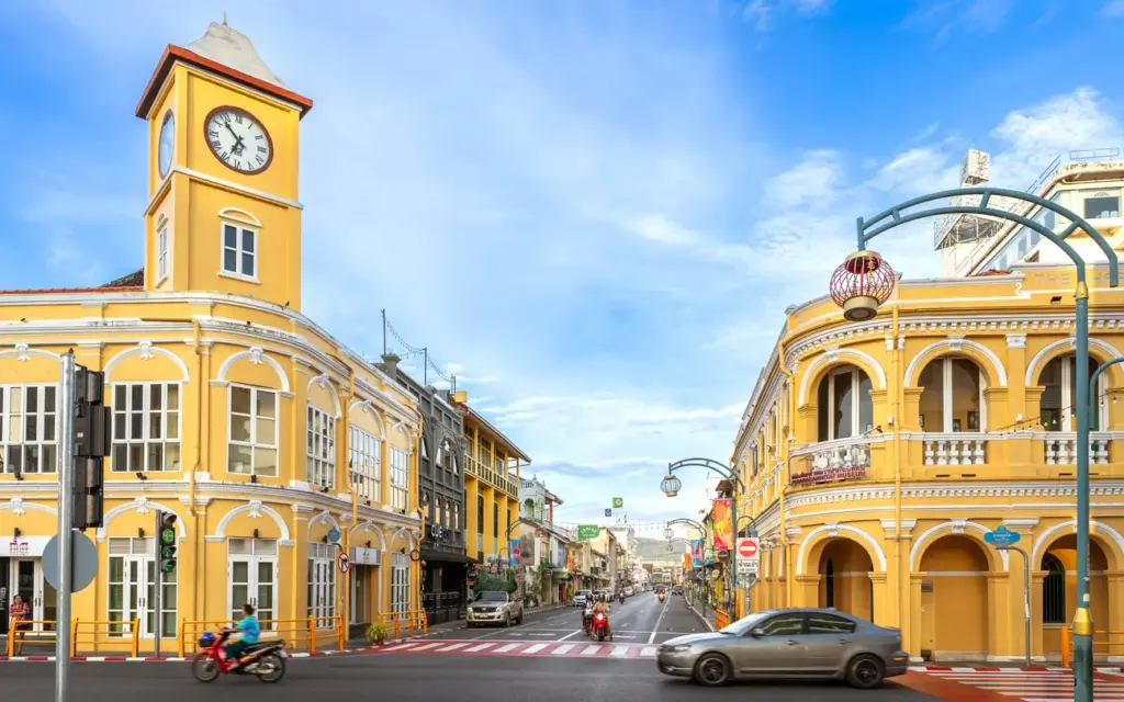 Phuket Eski Şehir'in canlı bir sokağında, geleneksel Sino-Portekiz mimarisinin sarı cepheli, beyaz işlemeli, kemerli pencereli ve balkonlu binaları gökyüzüne uzanıyor; sol tarafta belirgin bir saat kulesi ile süslenen bu tarihi doku, cadde üzerindeki araçlar ve zarif fenerlerle birlikte Phuket'te gezilecek yerlerin mistik atmosferini yansıtıyor.