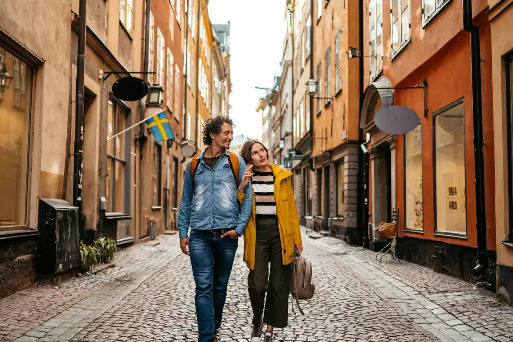 Kuzeyin En Rafine Rotası: Stockholm’de Gezilecek Yerler başlıklı içeriğimizle uyumlu olarak, tarihi Gamla Stan bölgesinin Arnavut kaldırımlı dar sokaklarından birinde, sarı yağmurluklu bir kadın ile kot gömlekli bir adamın merakla etrafı inceleyerek yürüdüğü, arka planda İsveç bayrağının asılı olduğu safran sarısı ve terra cotta renkli binaların görüldüğü canlı bir an.