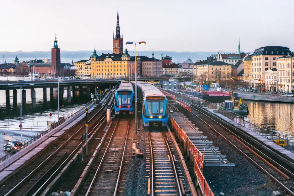 Stockholm'de iki modern mavi metro treni, Stadshuset ve tarihi yapıların yükseldiği şehrin panoramik siluetine doğru raylar üzerinde ilerleyerek, şehrin gelişmiş ulaşım ağının ve gezilecek yerlerin estetik entegrasyonunu vurguluyor.