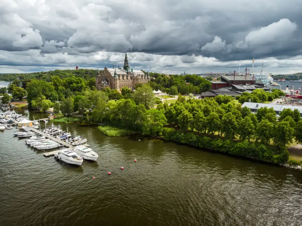 Gri bulutlu bir günde, Stockholm'ün yeşil Djurgården adasının kıyısında yer alan bir marinada sıralanmış beyaz tekneler, adanın kalbindeki görkemli Nordiska Museet ve kırmızı çatılı Vasa Müzesi'ni öne çıkararak, şehrin ikonik gezilecek yerlerini bütüncül bir bakışla sunuyor.