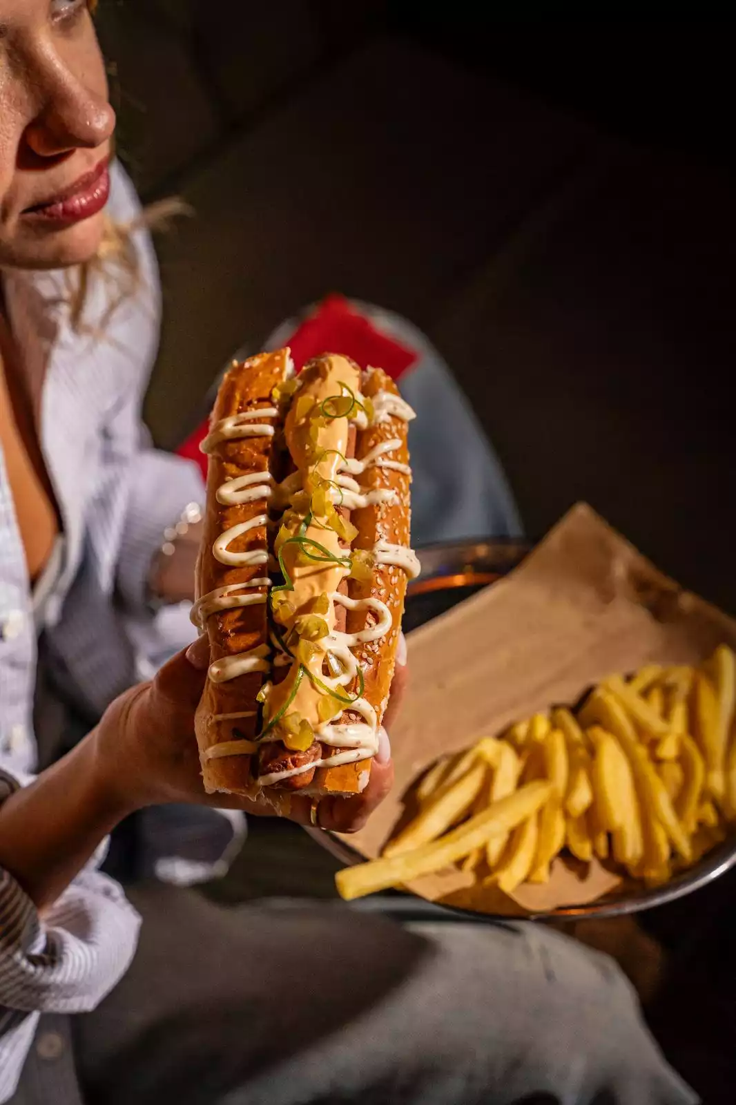 İstanbul’un: Oggusto İstanbul'un En İyi Hamburgercileri: Bir kadın, elinde hardal, mayonez ve jalapeno biberleriyle süslenmiş, susamlı ekmek arası gurme bir sosisli sandviç tutuyor. Arka planda metal tepsi ve kraft kağıt üzerinde servis edilen çıtır patates kızartmaları iştah kabartıyor.