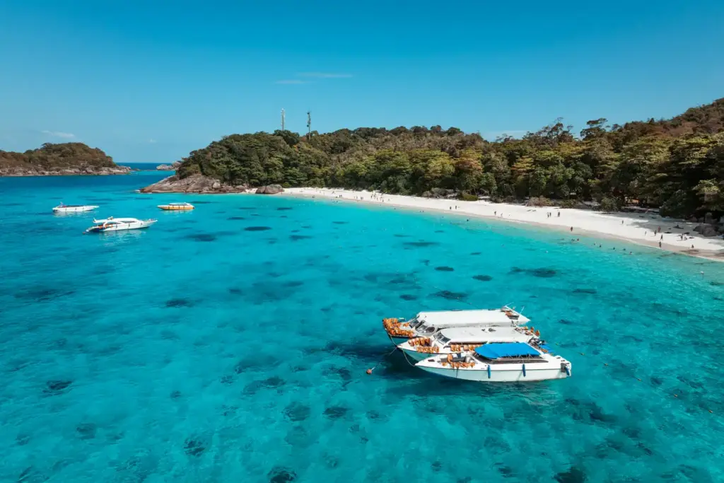 Phuket ve çevresindeki keşfedilmeyi bekleyen turkuaz kıyıların eşsiz güzelliğini gözler önüne seren bu geniş açılı görselde, masmavi berrak sular üzerinde demirlemiş birçok beyaz sürat teknesi, güneşlenen ve yüzen insanların olduğu bembeyaz bir kum plajı ile yemyeşil tropikal adalar göze çarpıyor.
