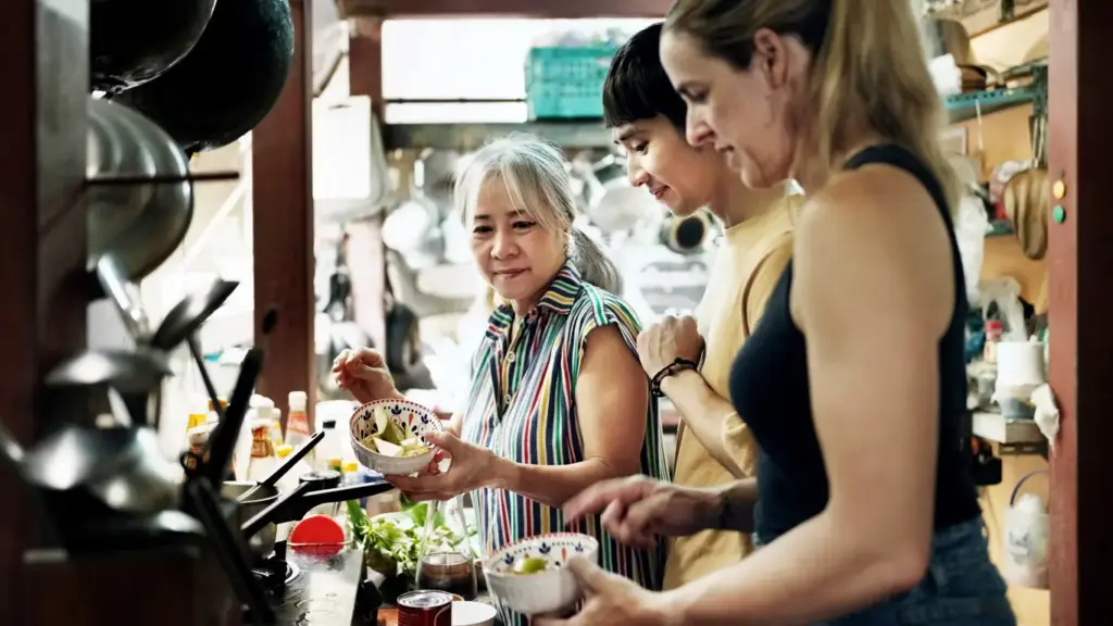 Phuket'in zengin gastronomi kültürünü deneyimlemek isteyen çeşitli kökenlerden gelen üç kişi, yaşlı bir Taylandlı kadının rehberliğinde geleneksel bir Thai yemek kursunda taze sebzeleri doğrayarak veya hazırlayarak aktif bir şekilde yemek yapıyor, otantik bir mutfağın sıcak atmosferinde keyifli anlar paylaşıyorlar.