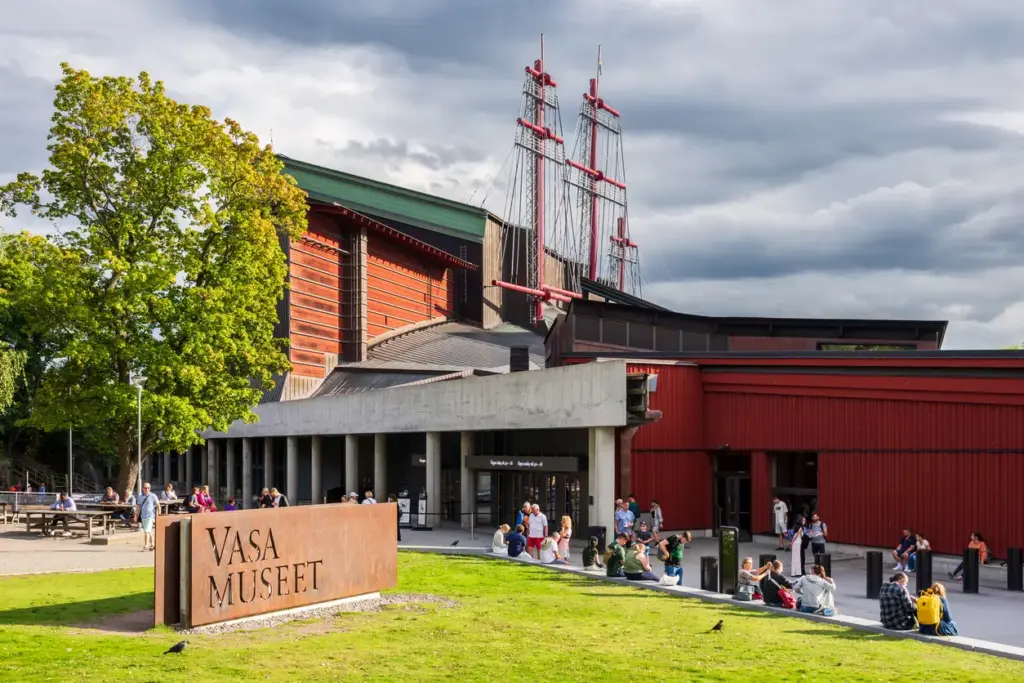 Stockholm'de mutlaka görülmesi gereken yerlerden Vasa Müzesi'nin kırmızı ve yeşil detaylı binası, çatısından yükselen devasa kırmızı gemi direkleri, önündeki VASA MUSEET yazılı paslı tabela ve yemyeşil çimenlikte oturan veya girişe yönelen çok sayıda ziyaretçiyle, şehrin kültürel zenginliğini ve Kuzeyin En Rafine Rotası rehberindeki önemini vurguluyor.