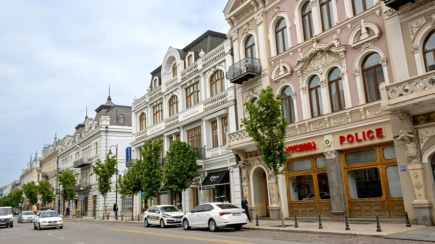 Bulutlu bir günde, Gürcistan'ın başkenti Tiflis'teki ünlü Agmashenebeli Caddesi boyunca sıralanmış, süslü cephelere ve zarif pencerelere sahip pastel tonlarda tarihi binalar, cadde üzerinde seyreden ve park etmiş araçlar ile yol kenarındaki genç ağaçların oluşturduğu canlı şehir manzarasını gösterir.