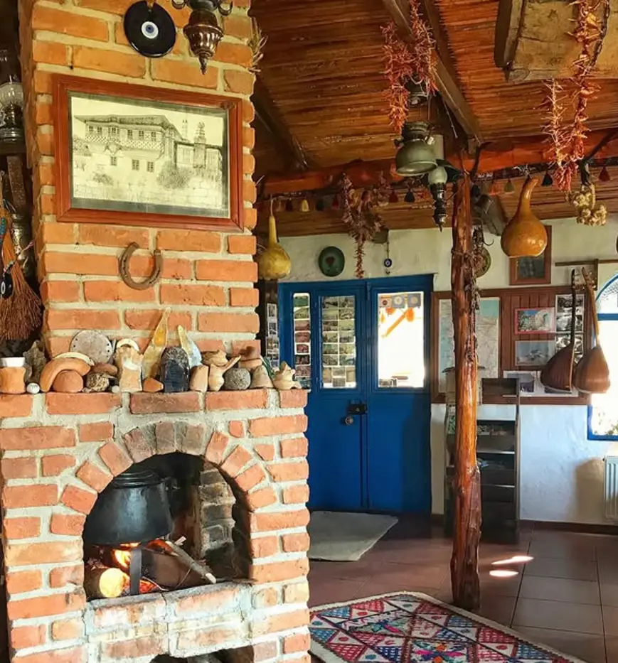 Sıcak ve davetkar bir pansiyon iç mekanı, ateşin yandığı tuğla bir şömine üzerinde büyük bir tencerenin bulunduğu, kuru kırmızı biberler ve kabaklarla süslenmiş ahşap tavan, arka planda parlak mavi çift kapı ve renkli kilimlerle dolu, geleneksel Agora Pansiyon konaklamasının samimi ve otantik bir atmosferini sergiliyor.