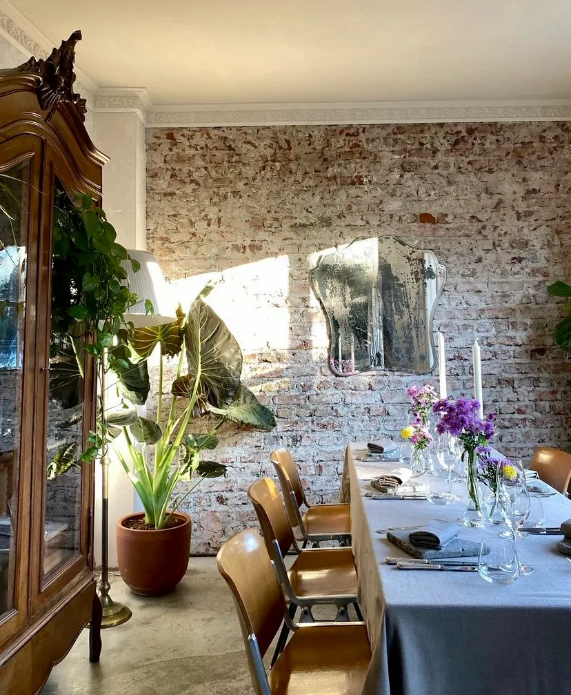 Araka restoranında güneşin vurduğu rustik tuğla duvarın önünde, mor ve sarı çiçek aranjmanları, zarif masa düzeni, ahşap sandalyeler, büyük yeşil bitkiler ve eski bir aynanın yansıdığı, misafirperver bir yemek alanı ambiyansı.