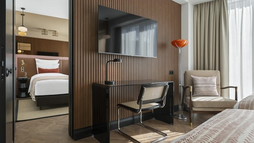 Tiflis'teki Artizan Design Hotel'in modern bir odasında, ahşap panelli duvara asılı düz ekran televizyonun altında şık bir çalışma masası ve hasır detaylı sandalye, sağda turuncu bir lambader ve krem rengi koltuk yer alırken, kapı aralığından yatak odası görülüyor.