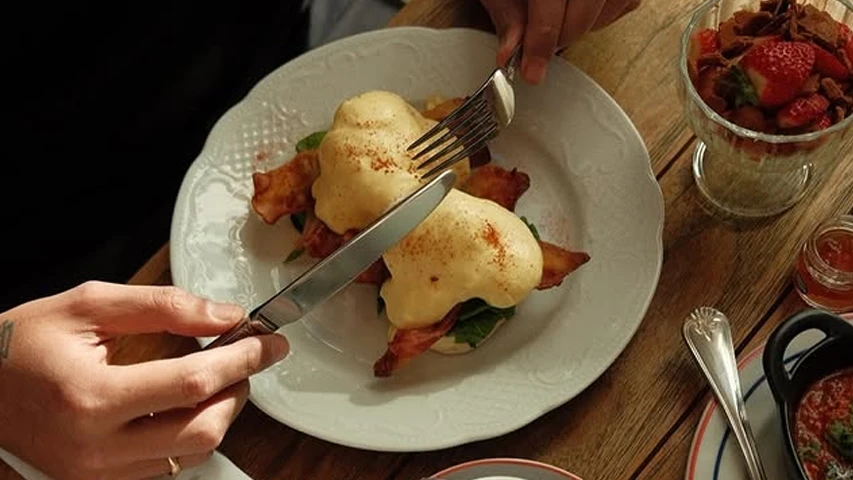 Tiflis'teki artizan restoranlardan birinde lezzetli bir öğün sunan bu görselde, bir kişi beyaz bir tabakta çıtır pastırma ve ıspanakla hazırlanan Yumurta Benedict'i keserken, yanında çilekli ve çikolatalı bir tatlı da görülüyor.