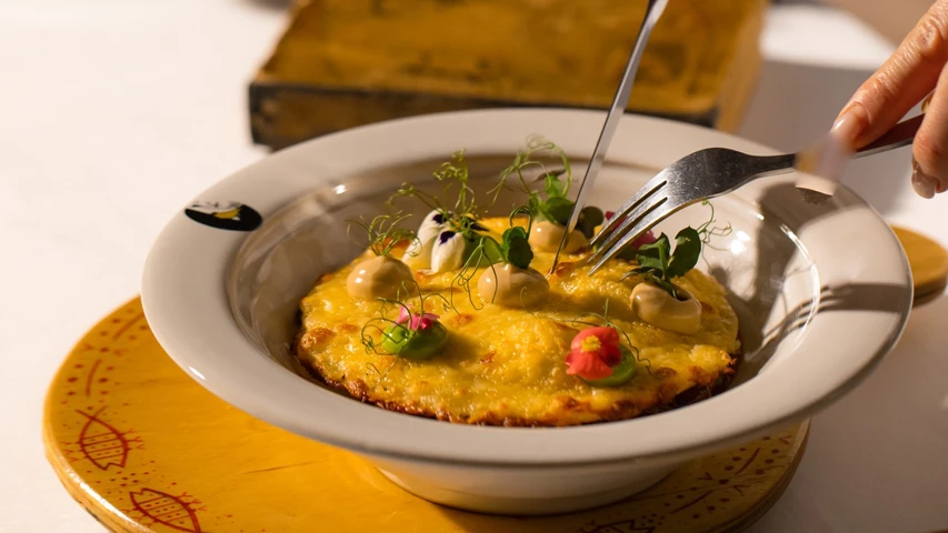 Barbarestan Tiflis restoranında servis edilen, üzerinde mikro yeşillikler, renkli yenilebilir çiçekler ve krem soslarla süslenmiş, altın sarısı renginde, fırınlanmış bir yemeğe çatal bıçakla uzanan bir elin yakın çekimi, Gürcü mutfağının modern ve zarif sunumunu vurguluyor.