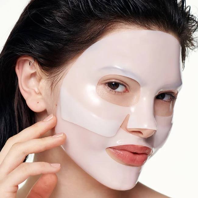 Şeffaf pembe renkli Biodance Real Deep Maske yüzüne özenle yerleştirilmiş, hafifçe ıslak görünen koyu saçlı bir kadın, maskeyi cildine nazikçe oturturken derinlemesine nemlendirme hissini yansıtan pürüzsüz ve ışıltılı bir cilde sahip.
