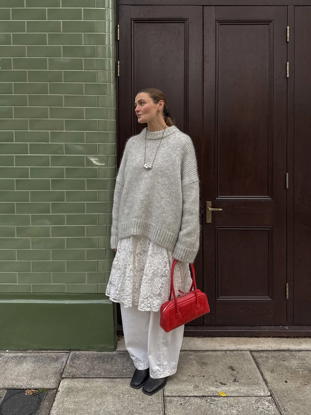 Gri oversize triko kazak, beyaz dantel elbise katmanı ve beyaz pantolonla oluşturulmuş modern günlük stil; kırmızı çanta detayıyla dantelin şehir stiline uyarlanışı.